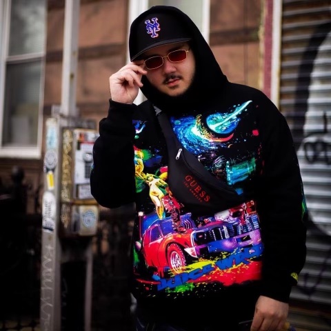 a light skinned man wearing a hoodie and sunglasses and baseball cap stands on the street