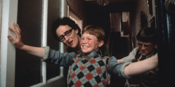 A red-headed boy in an argyle vest smilingly holds his arms across a doorway as two others peek out behind him