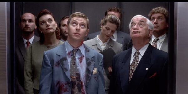A man in a bloodied suit stands in an elevator and looks up, surrounded by others