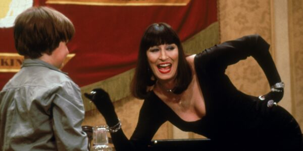 A smiling woman in a black wig leans on a table with a little boy next to her