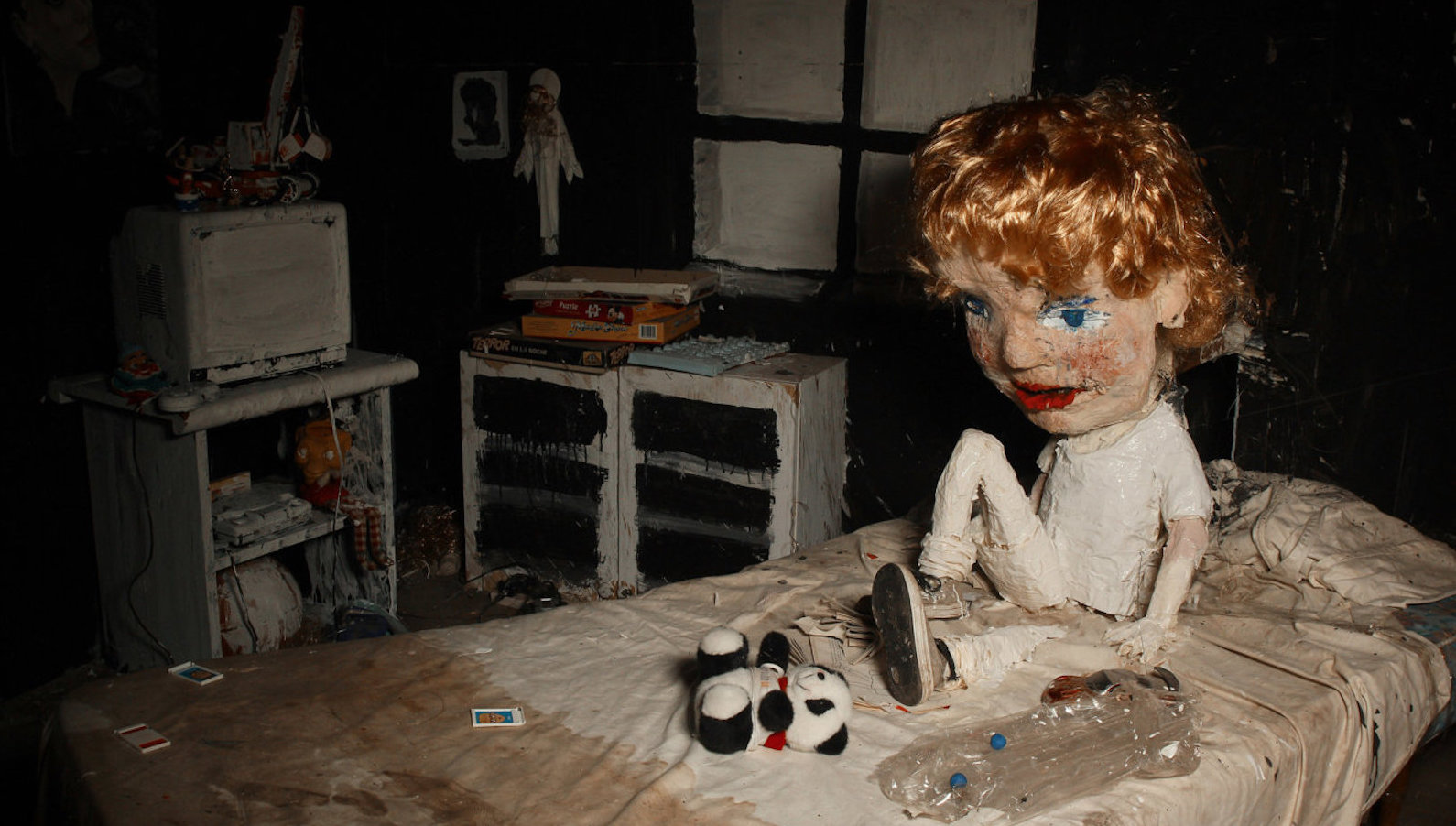 A melancholy puppet with red hair sits on a dirty bed with a toy panda bear