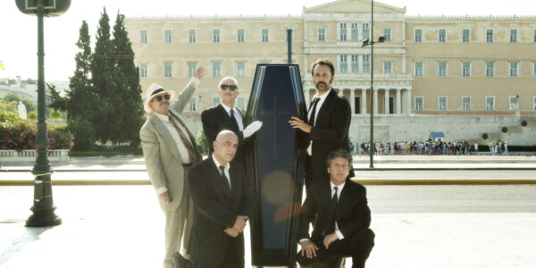 Five men in suits stand around an upright closed coffin on a city street in front of a municipal building