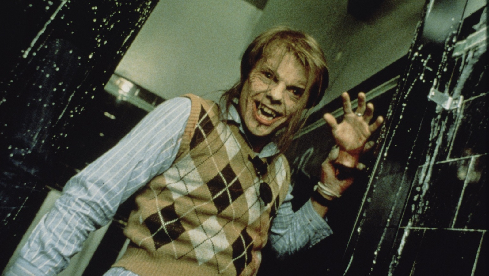 A man in an argyle vest and wearing fangs holds up a severed hand and smiles creepily at the camera