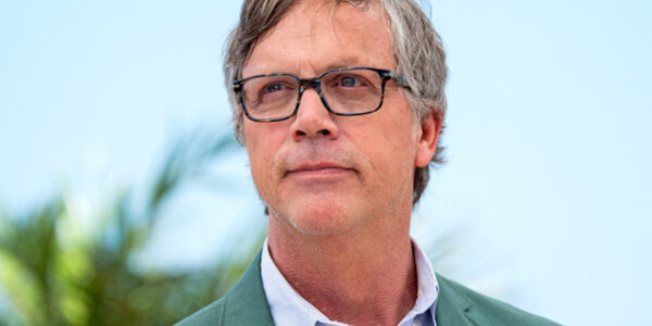 A white man wearing glasses, a blue shirt and green jacket, seen in a head shot against a blue sky
