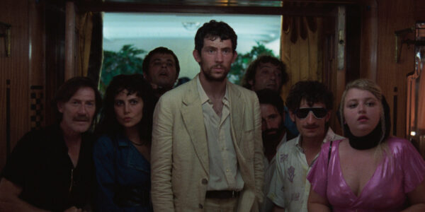 A man in a rumpled white suit surrounded by seven shorter people stands in a room near a window, all of them looking interested past the camera