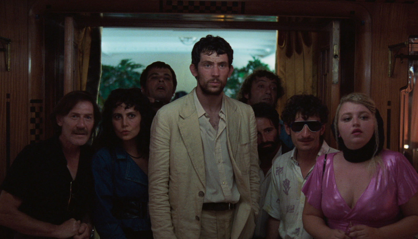 A man in a rumpled white suit surrounded by seven shorter people stands in a room near a window, all of them looking interested past the camera