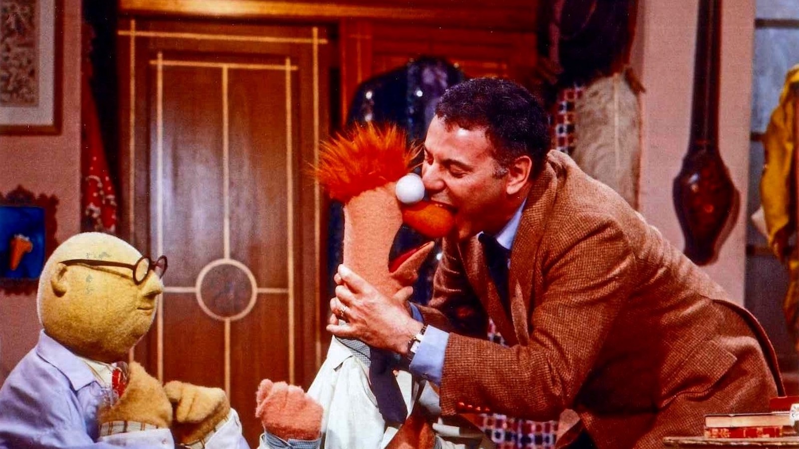 A man in a brown blazer bites a muppet on its red nose while another muppet with glasses watches.