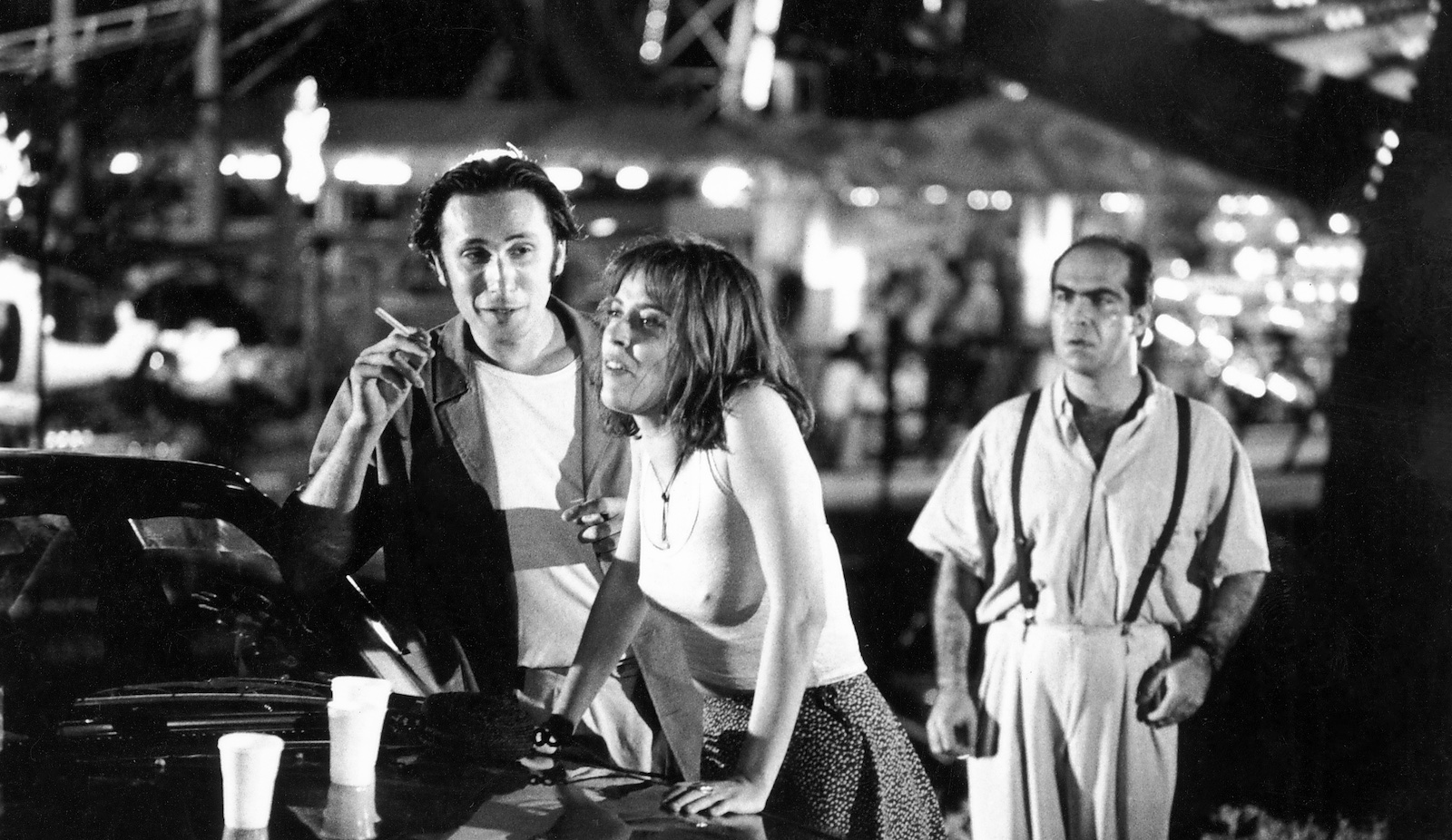 NO BUDGET STORY-006 Three people stand by a car in an outdoor carnival, a woman leaning against the car hood and a man holding a cigarette talking to her, while another man stands behind them and looks at them