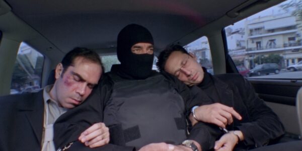 Three men sit in the back of a car, handcuffed to each other, the middle man wearing a bulletproof vest and ski mask