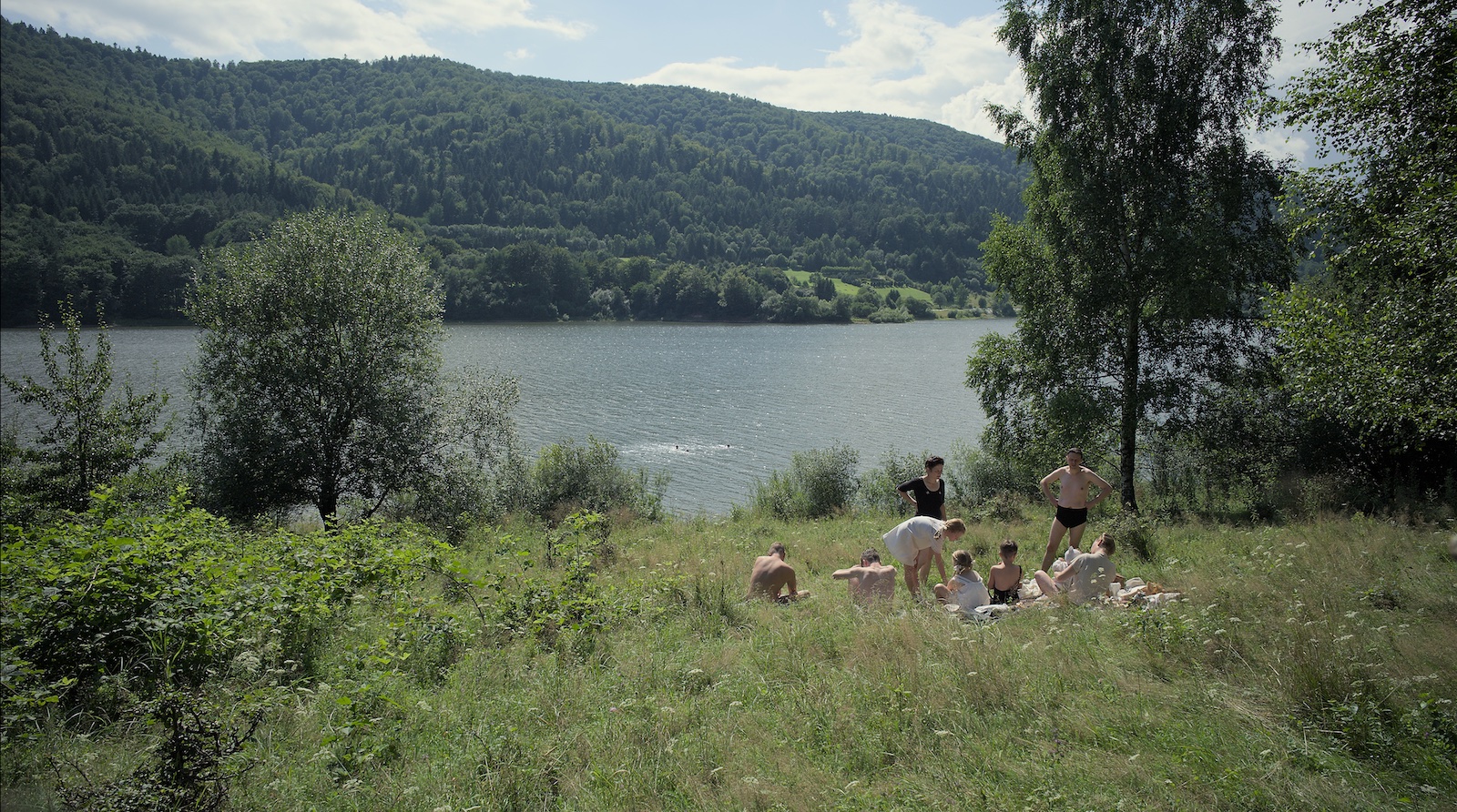 A group of people, children and adults, in bathing suits by a beautiful idyllic lake, sitting in the grass