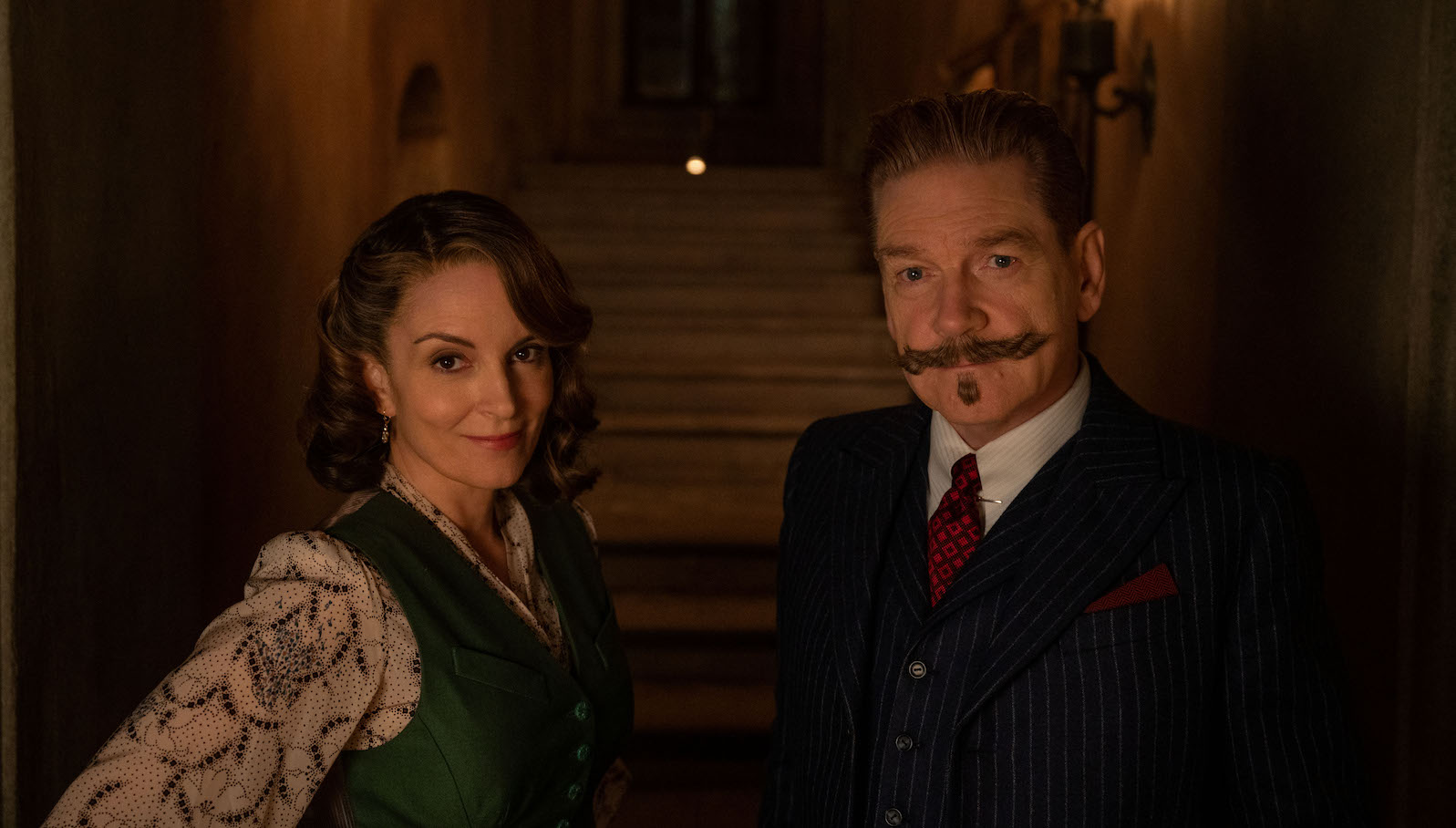 A woman in a green vest and a man wth a twirly mustache look into the camera.