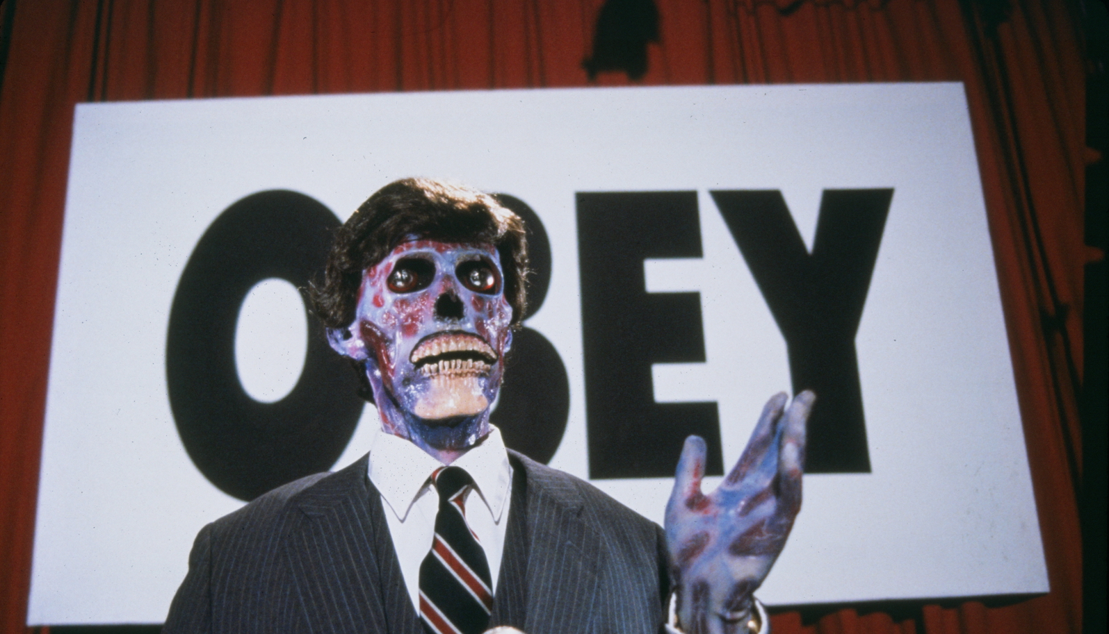 theylive A suited man with a skeleton face stands before a large sign that says OBEY.