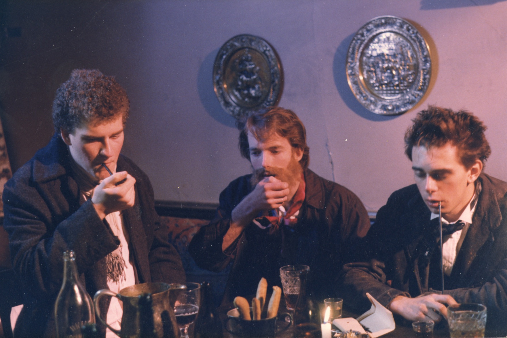 Three men in old-fashioned clothes sit at a table smoking pipes
