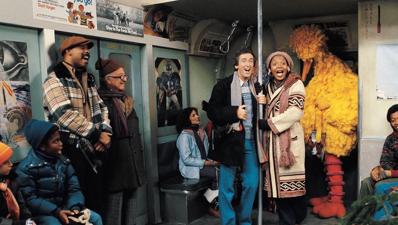 Two people hold a subway pole while singing, and a big yellow muppet bird stands behind them.