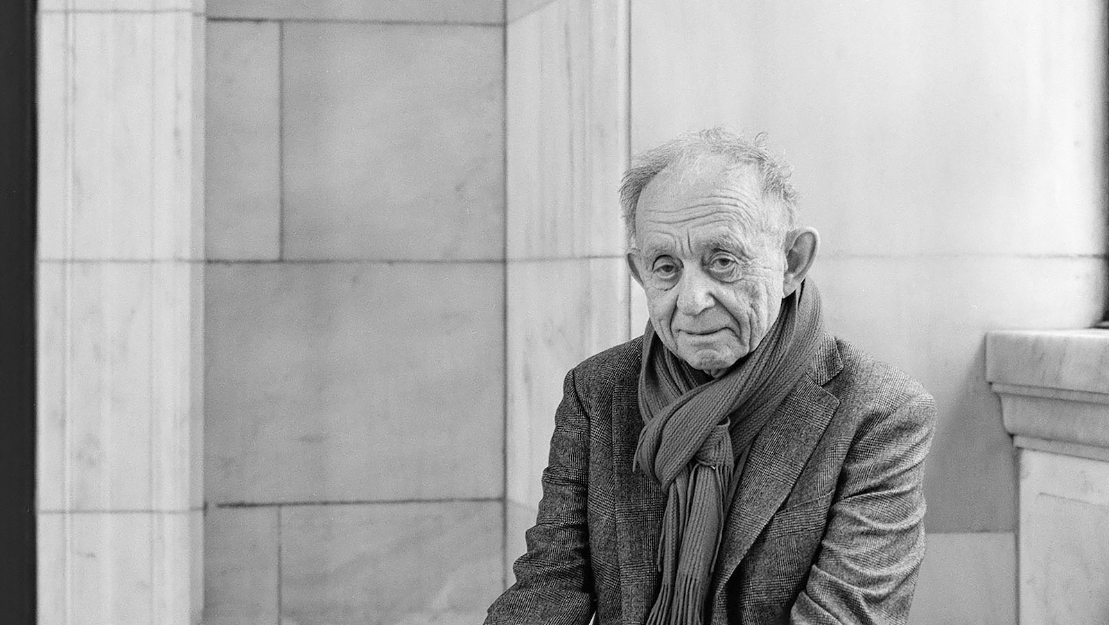 a black and white image of a man looking at the camera wearing a scarf, sitting outside a library wall