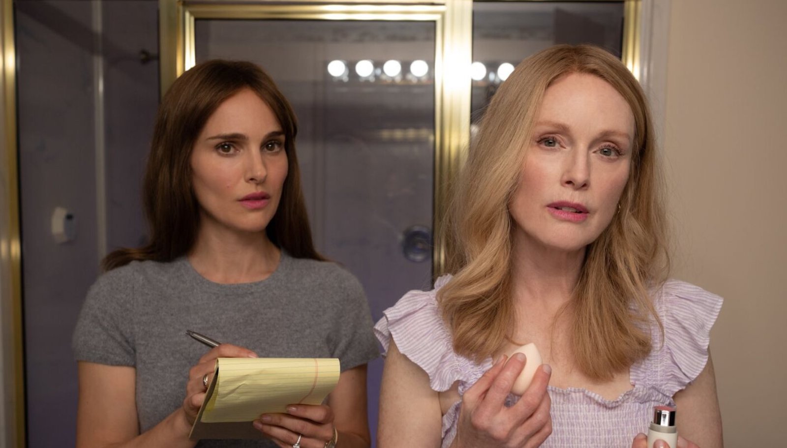 Two women look in a mirror, one holding makeup, one holding a pad of paper