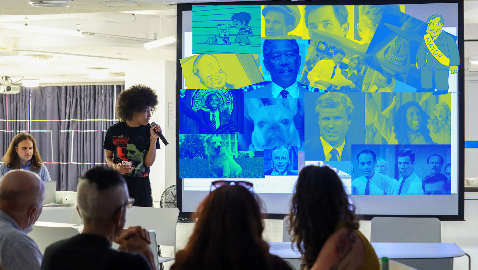 A woman holds a microphone and talks in front of an audience and standing before a screen made up of blue and yellow images of actors