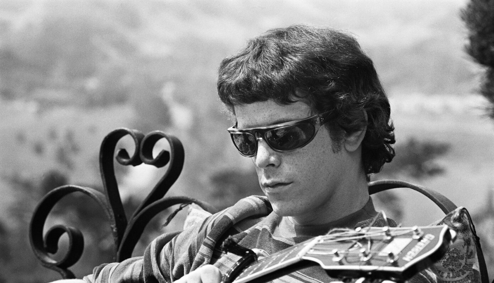 A man in sunglasses holds a guitar sitting outside