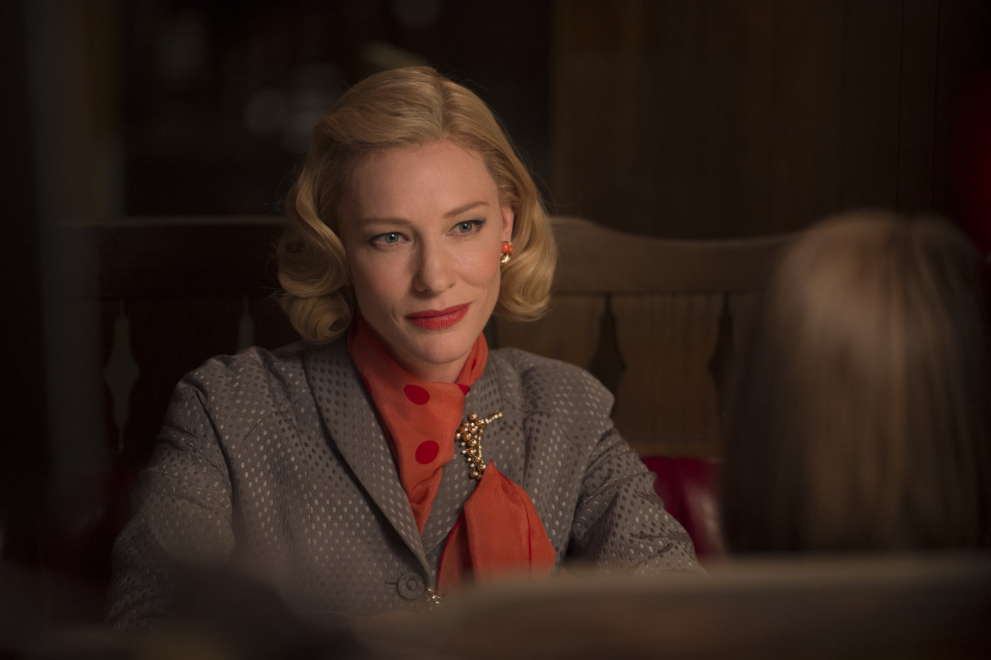 A woman with coiffed hair and a red scarf sits at a restaurant and gazes lovingly across a table toward camera