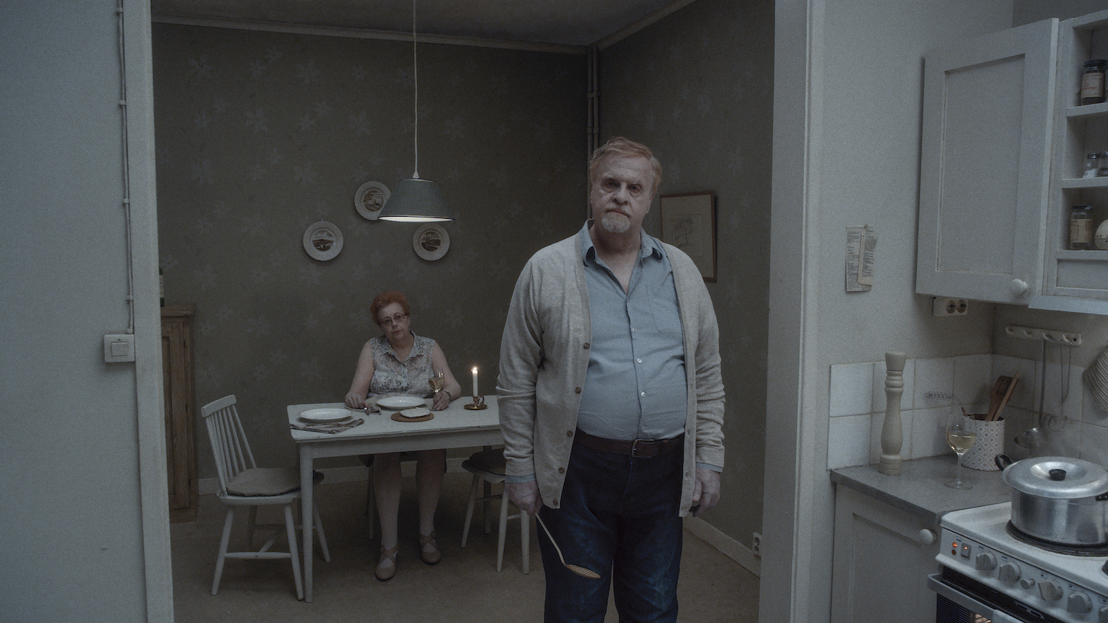 A man stands in a grim gray kitchen holding a spoon and staring past camera while a woman sits at a dinner table behind him