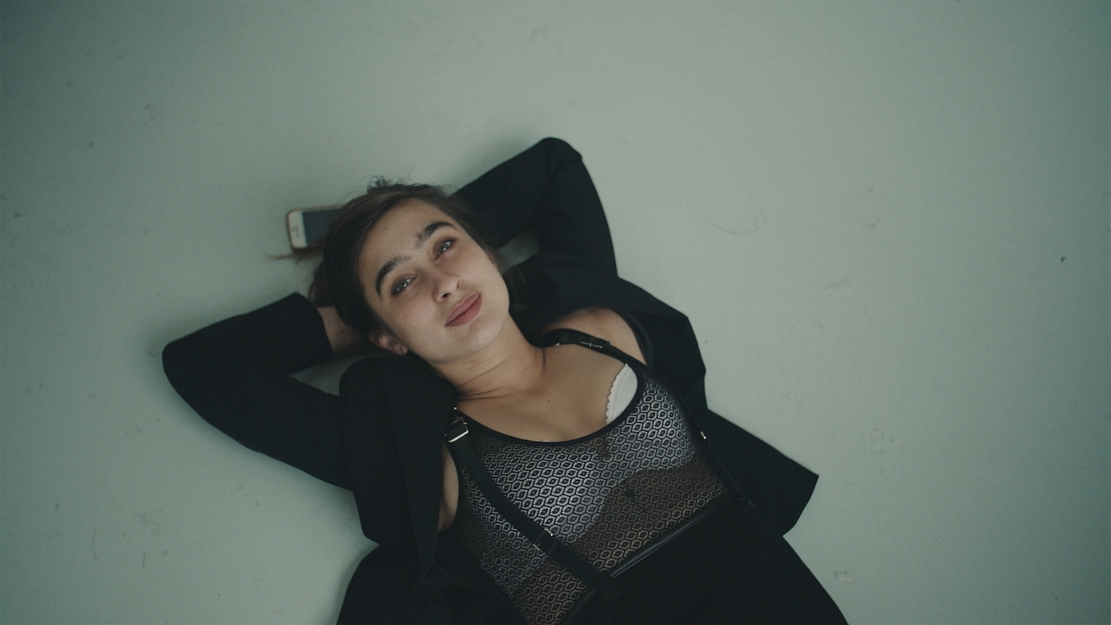 A woman lies on the floor with her hands behind her head, looking up and smiling, her cell phone behind her head.