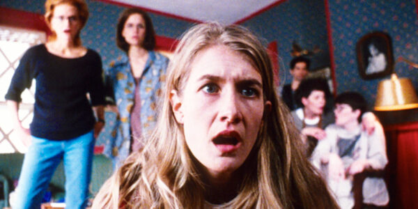 A woman stares slackjawed into the camera, crouching on the floor at TV level, with other women behind her standing also in shock