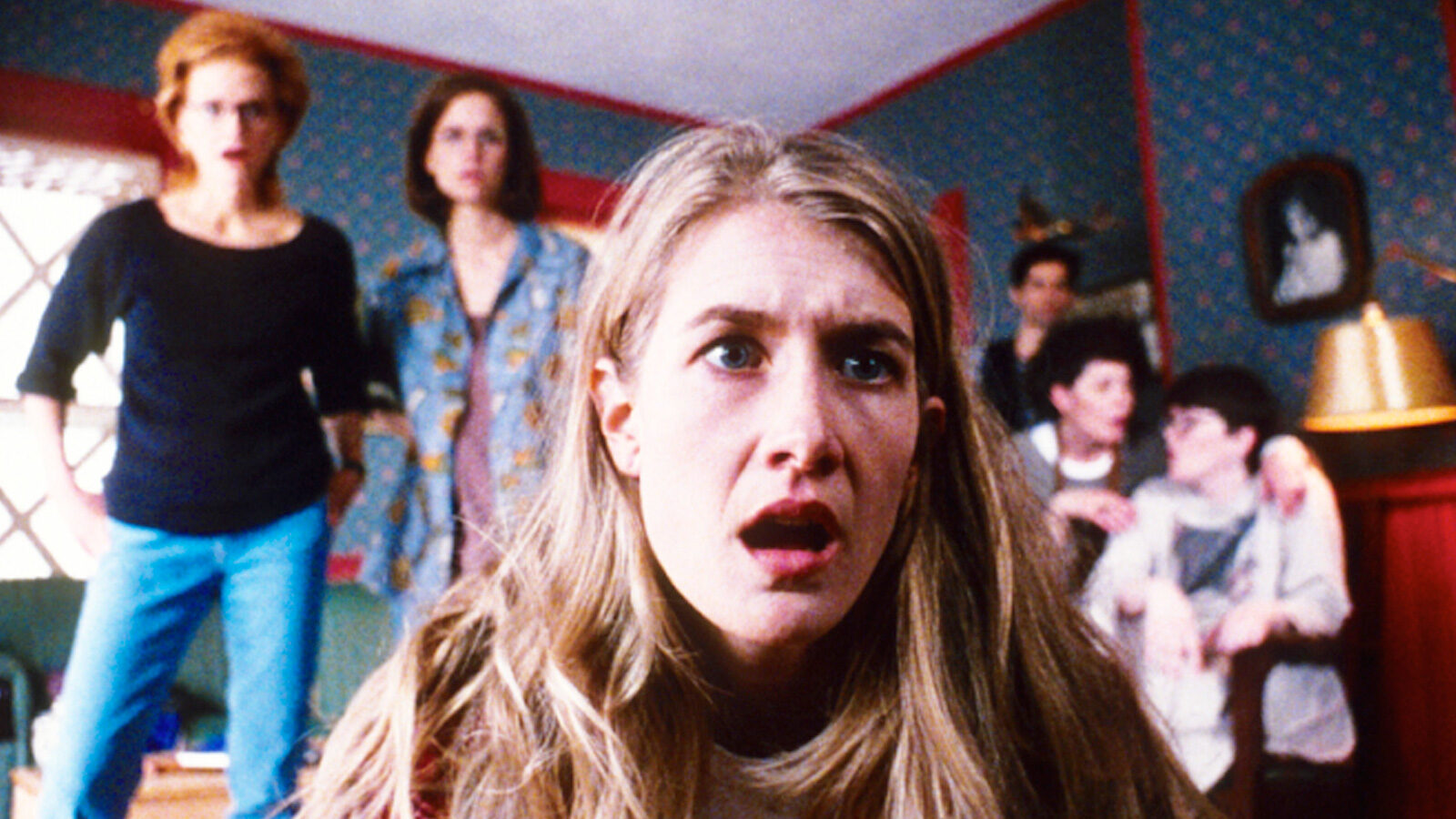 A woman stares slackjawed into the camera, crouching on the floor at TV level, with other women behind her standing also in shock