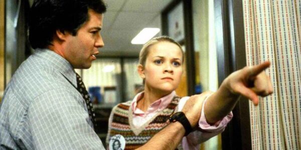 A man holds back a young woman as she aggressively points at someone off screen with a mean look on her face