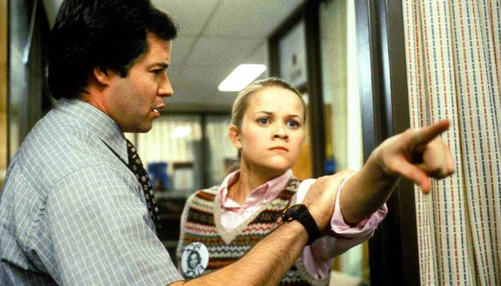 A man holds back a young woman as she aggressively points at someone off screen with a mean look on her face