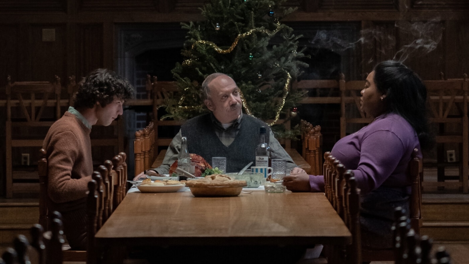 Three people sit around a dinner table talking to each other and eating with a Christmas tree behind the man who sits at the head of the table