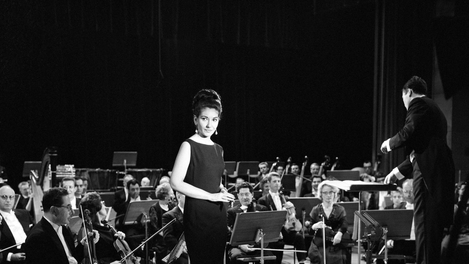 A regal singer in a black dress stands in a symphony space. She is surrounded by an orchestra.