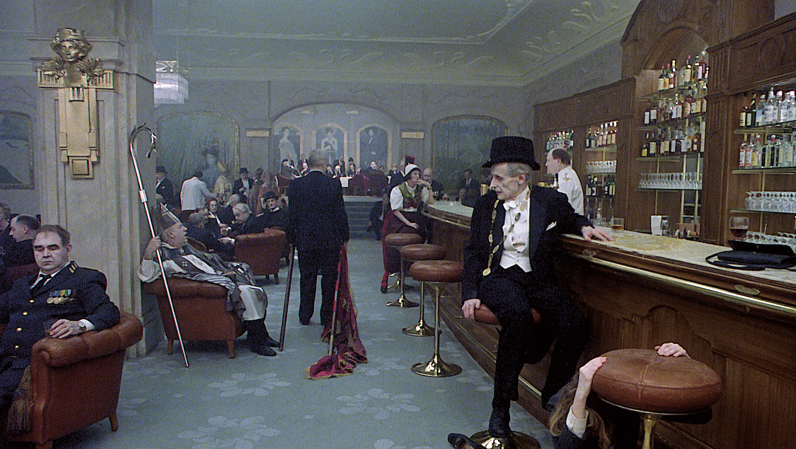 An old man in a top hat sits at a bar in a gray, smokey room.
