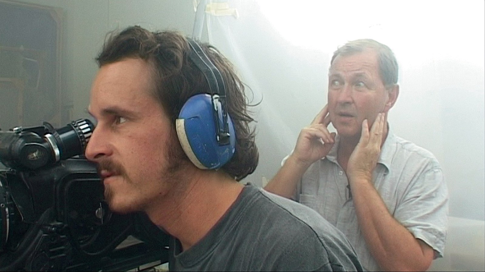 A man holding his fingers in his ears stands behind a man wearing blue sound-canceling headphones