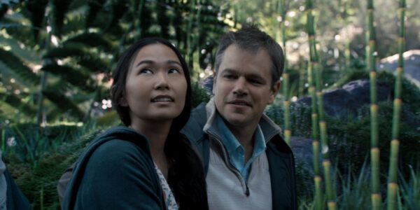 A man and woman walk through a green forest area, both smiling; she looks away and up to the sky with wonder, he looks at her.