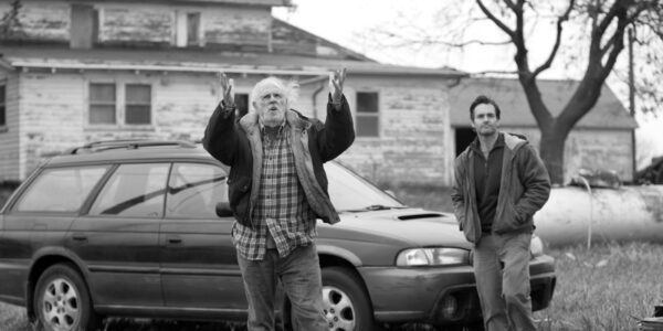 A black and white image of a disheveled man raising his hands to the sky, standing in front of a car, with a younger man standing to his left