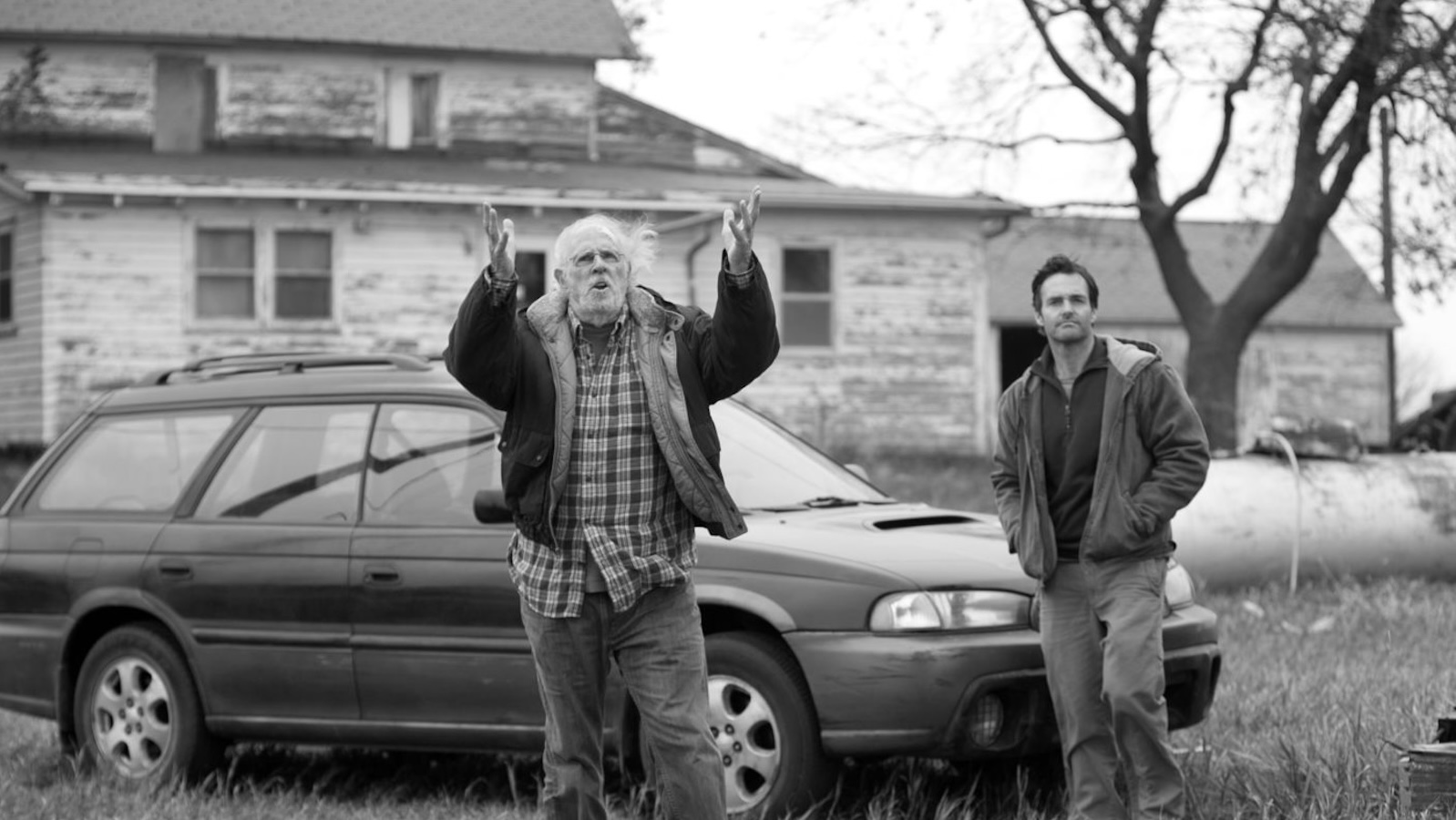 A black and white image of a disheveled man raising his hands to the sky, standing in front of a car, with a younger man standing to his left