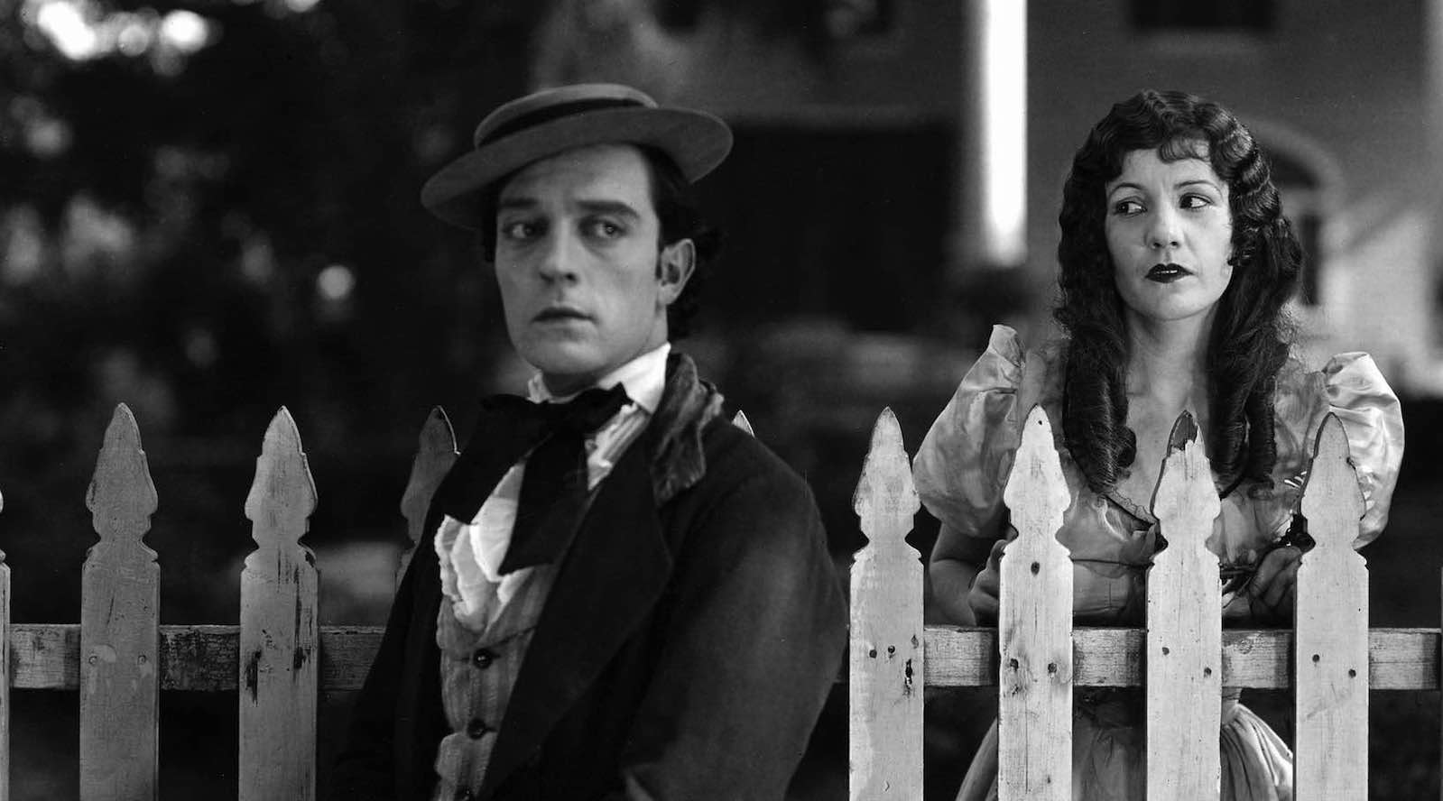 A man clutching a hat in his hand stands in front of a white picket fence, with a woman standing behind him behind the fence