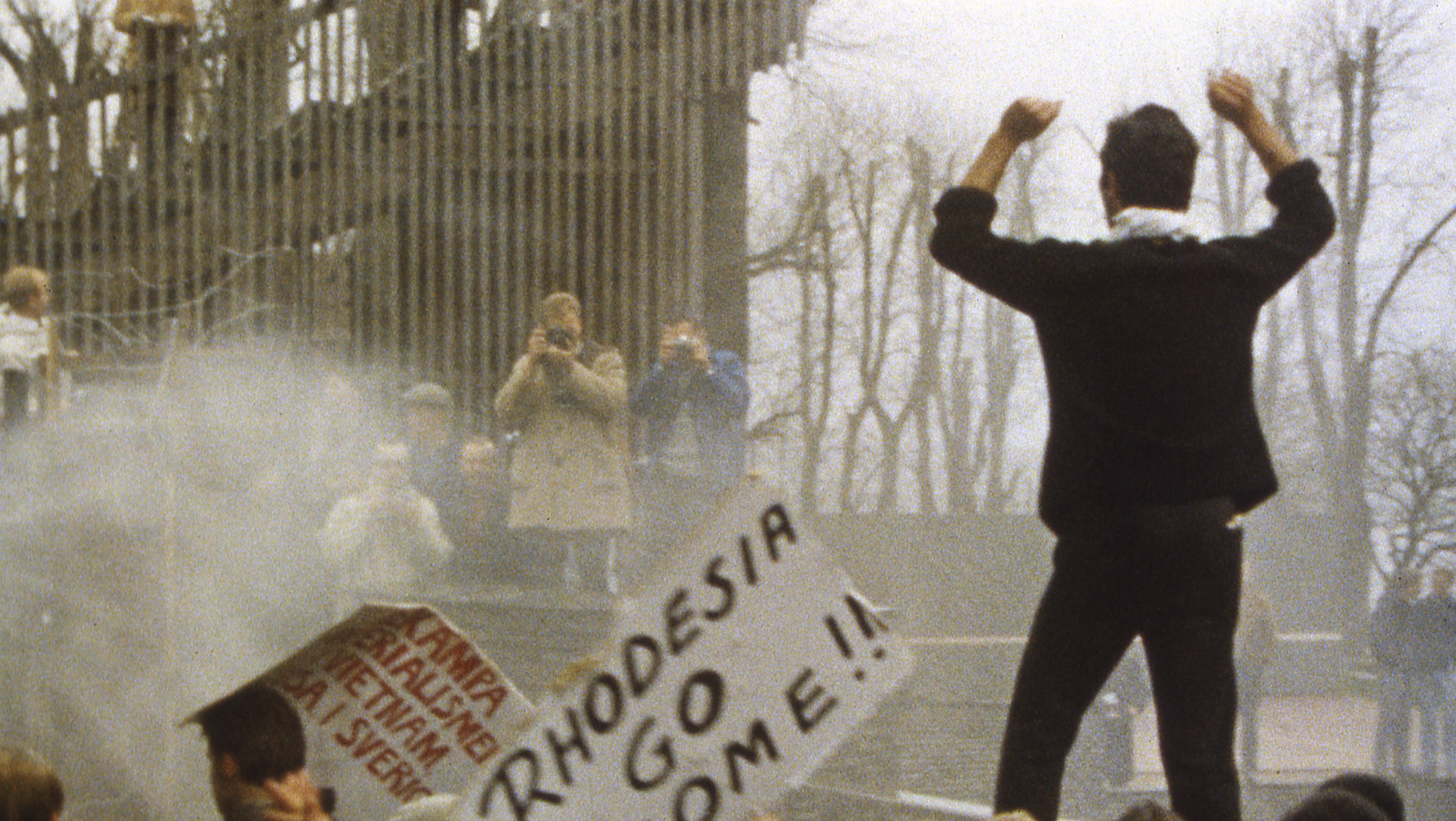 A man stands at a street protest with his arms in the air while people in the distance take photos of him; a sign near him says RHODESIA GO HOME!