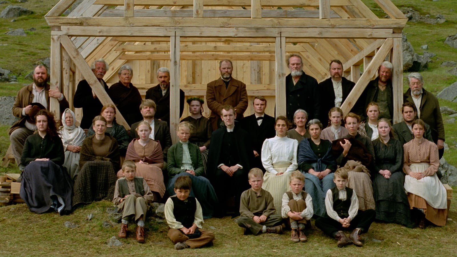 A large family dressed in 19th century clothes stands in front of a house being built amidst a rocky, green landscape, as though posing for a photo