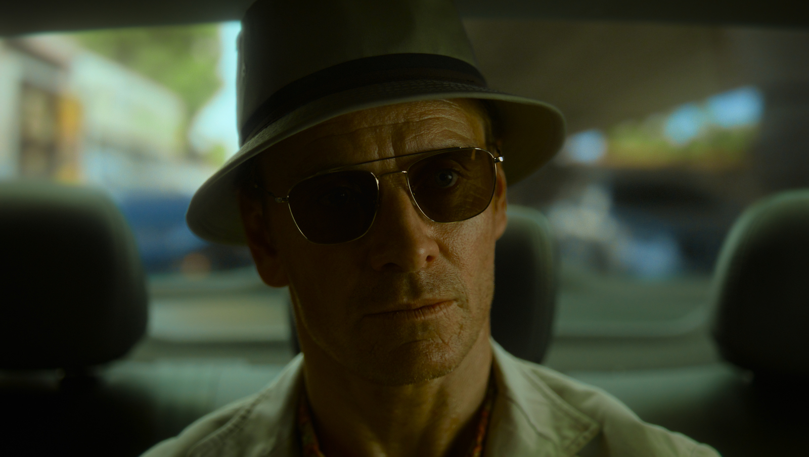 A man in sunglasses and a fedora sits in the back of a car looking straight ahead with a serious expression