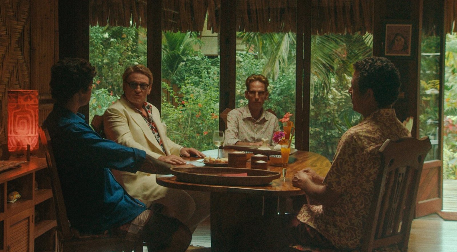 Four men sit around a table in tropical-colored clothes with dense green jungle leaves outside the window