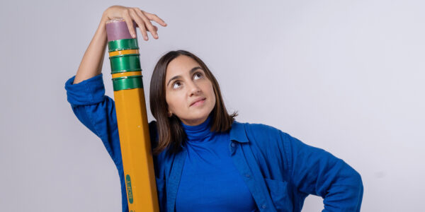 A woman stands against a white wall wearing a blue short and holding an enormous yellow pencil