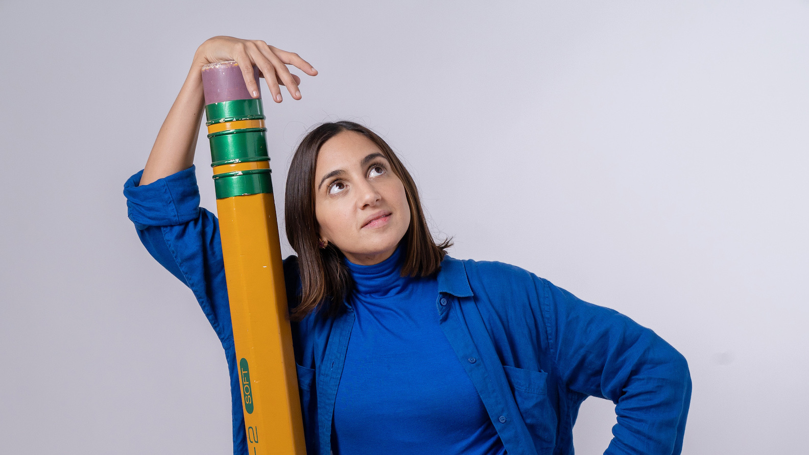 A woman stands against a white wall wearing a blue short and holding an enormous yellow pencil