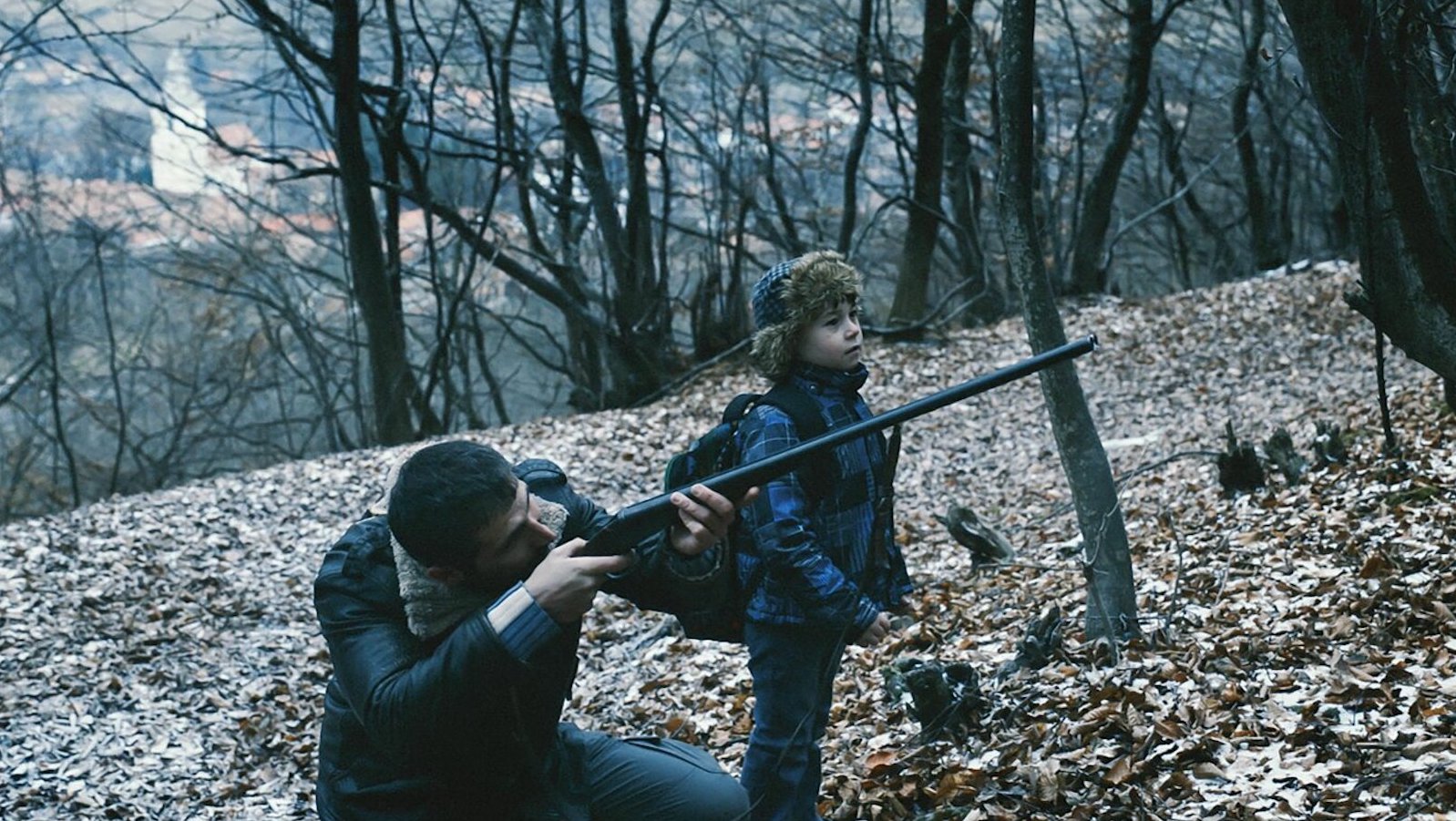 A man and a boy in the woods, surrounded by bare trees and leaves on the ground; the man aims a rifle off screen