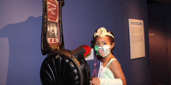 A little girl in a white dress and facemask looks at camera while standing next to a mutoscope device at Museum of the Moving Image