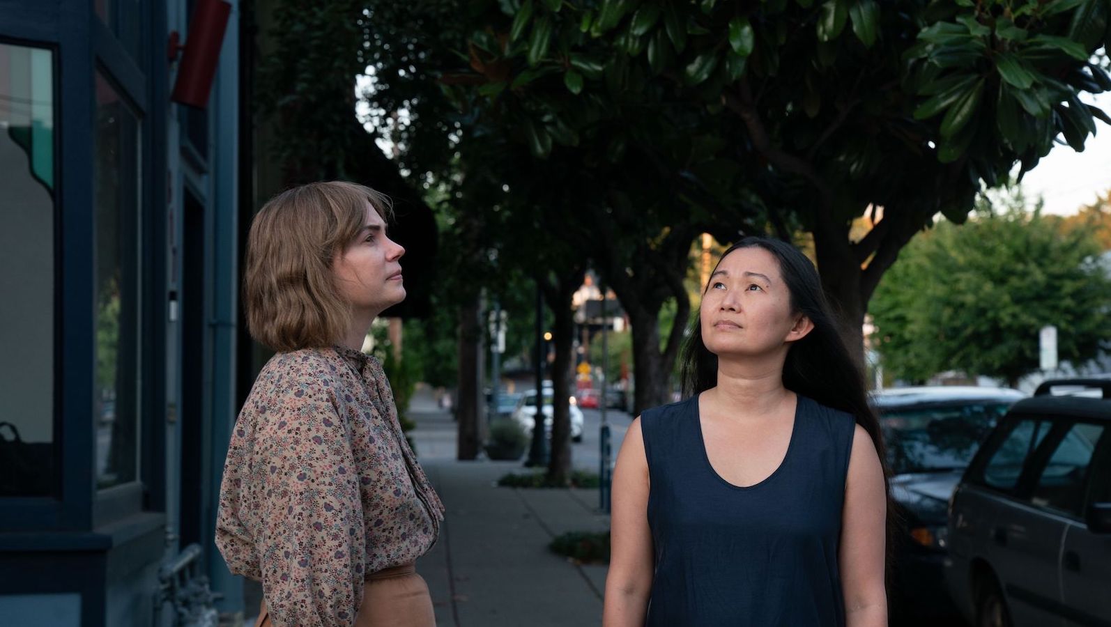 Two women stand on a city street in the afternoon sun and look up to the sky in different directions