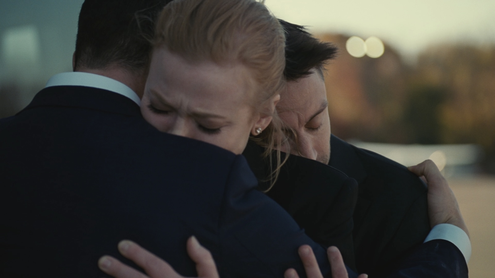 Three people hug each other with eyes closed in dark clothes, a woman in between two men