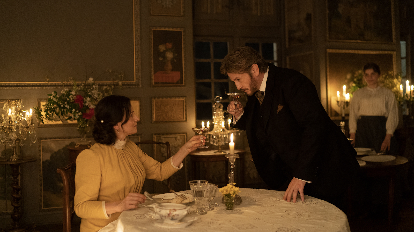 In an old-fashioned candlelit dining room, a man bows over a seated woman, both holding delicate cocktail glasses.