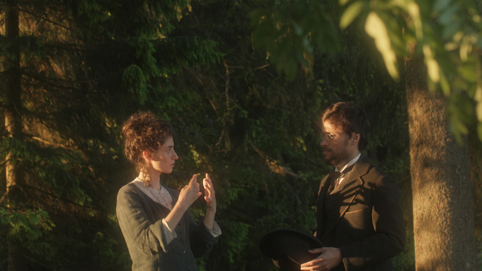 A woman and man in 19th century clothes talk to each other outside amidst a green forest setting