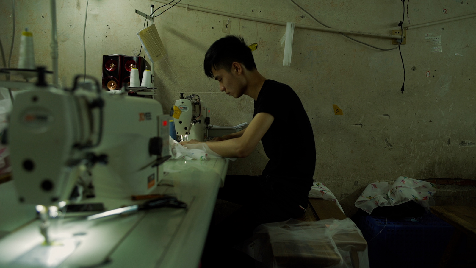 A young man sits over a sewing machine in a drab clothing factory